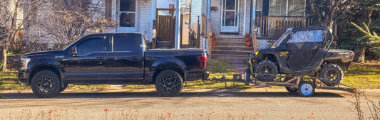 A black Ford F-150 truck towing a Can-Am Maverick X3 UTV on a trailer, parked in front of a white house with Halloween pumpkins on the porch steps in a suburban neighborhood during autumn.