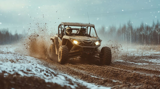 A muddy UTV with two helmeted riders driving through a snowy, dirt-covered field, kicking up dust and snow under a clear sky with trees in the background.