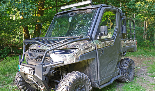 A utility vehicle with a Can-Am windshield parked amidst trees in a serene woodland setting.