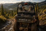 Two riders in an Arctic Cat Prowler side-by-side, shown from the rear on a muddy mountain trail. TerraRider rear UTV windshields available for 2006–2010 models.