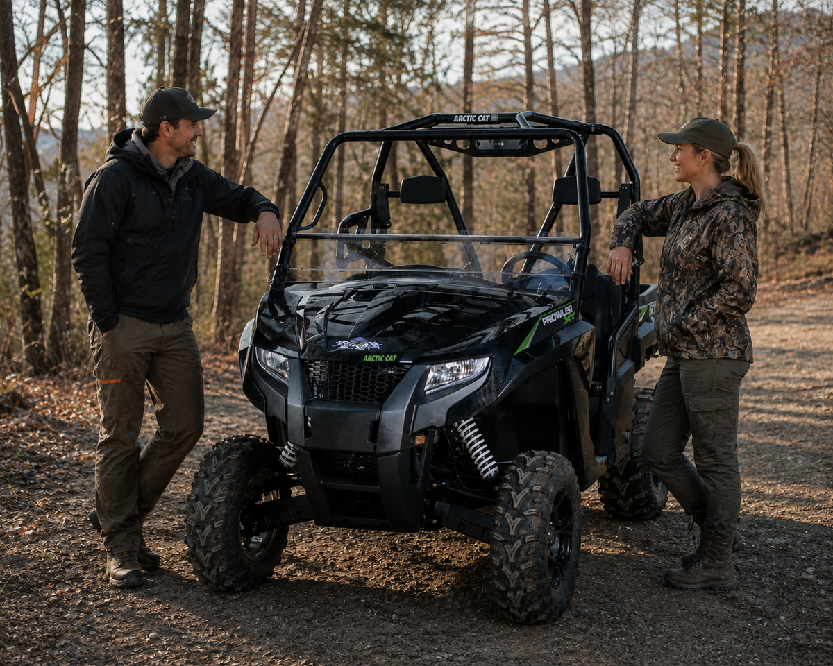 Two riders standing beside a black Arctic Cat Prowler with tilting windshield on a wooded trail