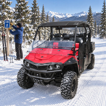 Red Mahindra mPACT XTV with tilting windshield on packed snow among evergreens, driver standing nearby. gallery-1