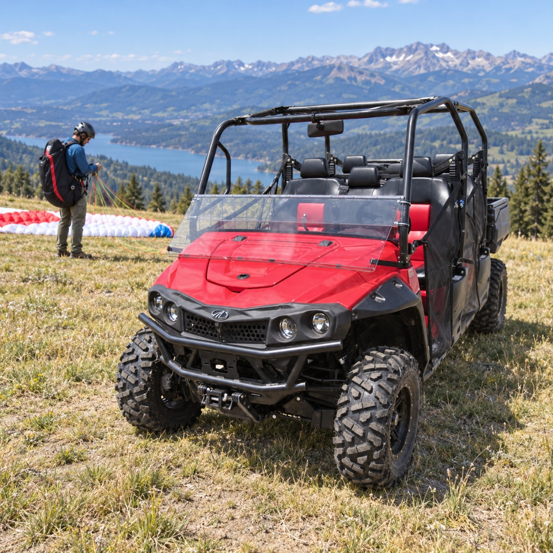 Red Mahindra mPACT XTV with tilting windshield on an alpine meadow above a lake, rider standing alongside. gallery-2