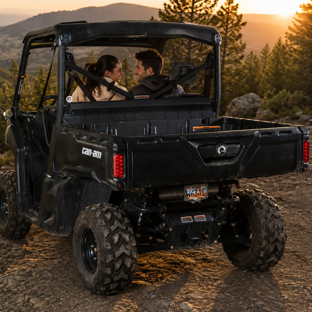 Two riders in a black Can-Am Defender with back windshield on a forest trail at sunset