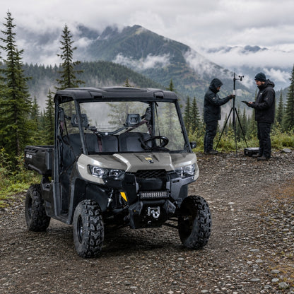Tan Can-Am Defender with half windshield on a trail with two people alongside