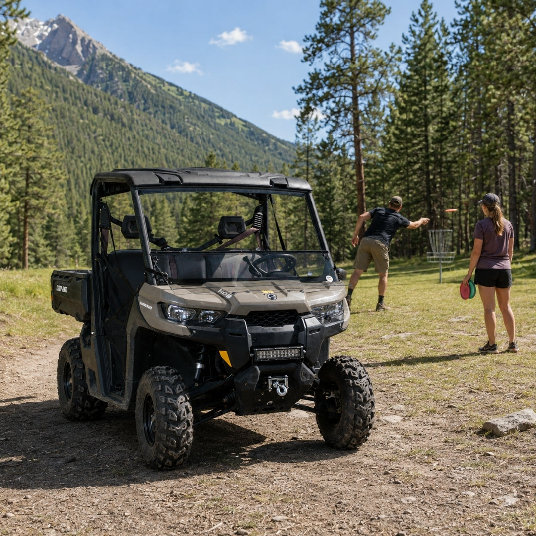 Tan Can-Am Defender with tilting windshield parked in a meadow with people alongside