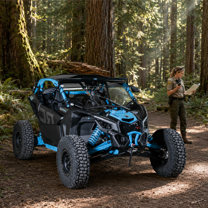 Blue Maverick X3 with full windshield parked among tall redwoods, rider standing alongside. gallery-2