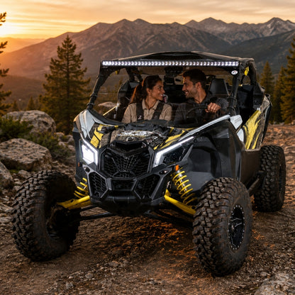 Two riders in a Maverick X3 with half windshield cresting a rock ledge at sunset