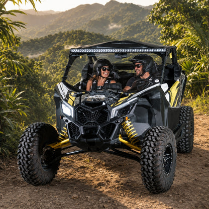 Black and yellow Maverick X3 with full windshield on a grassy mountain track, riders aboard. gallery-1