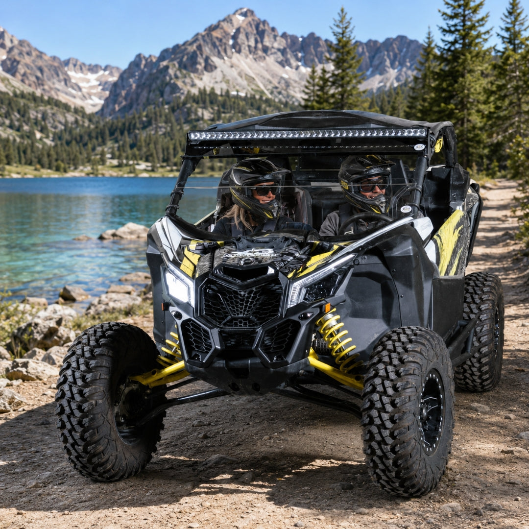 Maverick X3 with half windshield parked on a lakeshore below granite peaks, riders aboard