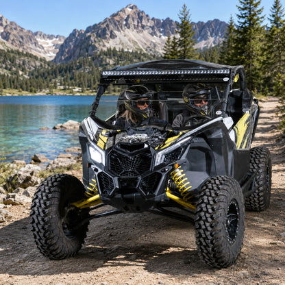 Maverick X3 with half windshield parked on a lakeshore below granite peaks, riders aboard